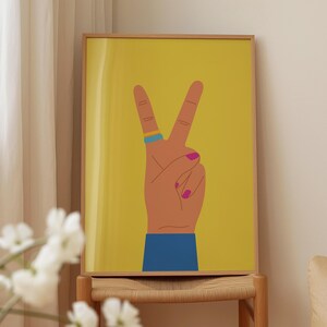 May include: A framed art print featuring a hand making the peace sign against a yellow background. The hand has a tan skin tone, pink fingernails, and a blue and yellow ring. The sleeve is blue.
