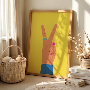 May include: A framed art print featuring a hand making the peace sign against a yellow background. The hand has a blue ring, pink fingernails, and a blue sleeve. The frame is made of wood. A wicker basket with stuffed toys sits nearby.