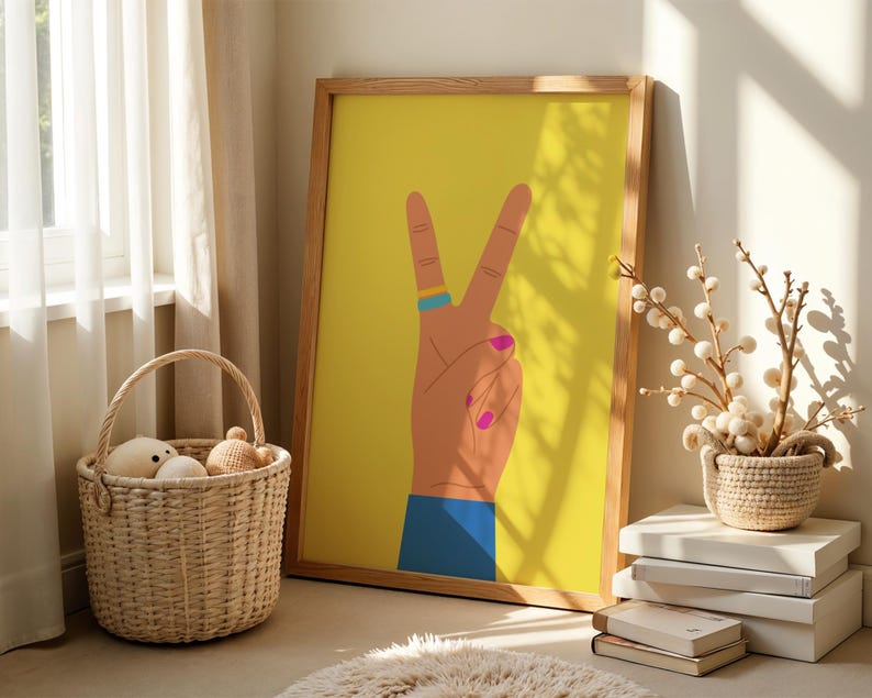 May include: A framed art print featuring a hand making the peace sign against a yellow background. The hand has a blue ring, pink fingernails, and a blue sleeve. The frame is made of wood. A wicker basket with stuffed toys sits nearby.