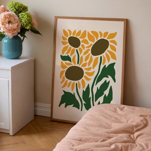 May include: A framed art print featuring a stylized illustration of three sunflowers. The sunflowers have yellow petals and brown centers, with green stems and leaves against a cream background. The frame is a light brown color.