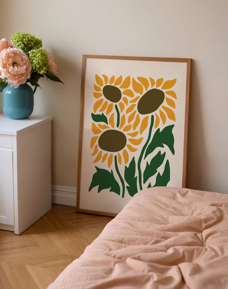 May include: A framed art print featuring a stylized illustration of three sunflowers. The sunflowers have yellow petals and brown centers, with green stems and leaves against a cream background. The frame is a light brown color.