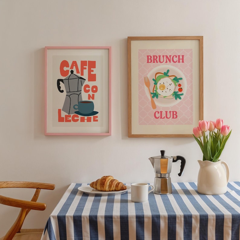 May include: Two framed art prints on a white wall. One pink-framed print says "CAFE CON LECHE" with a coffee pot and cup illustration. The other, in a wooden frame, says "BRUNCH CLUB" with a plate of food. A table with a blue and white striped cloth is in front.