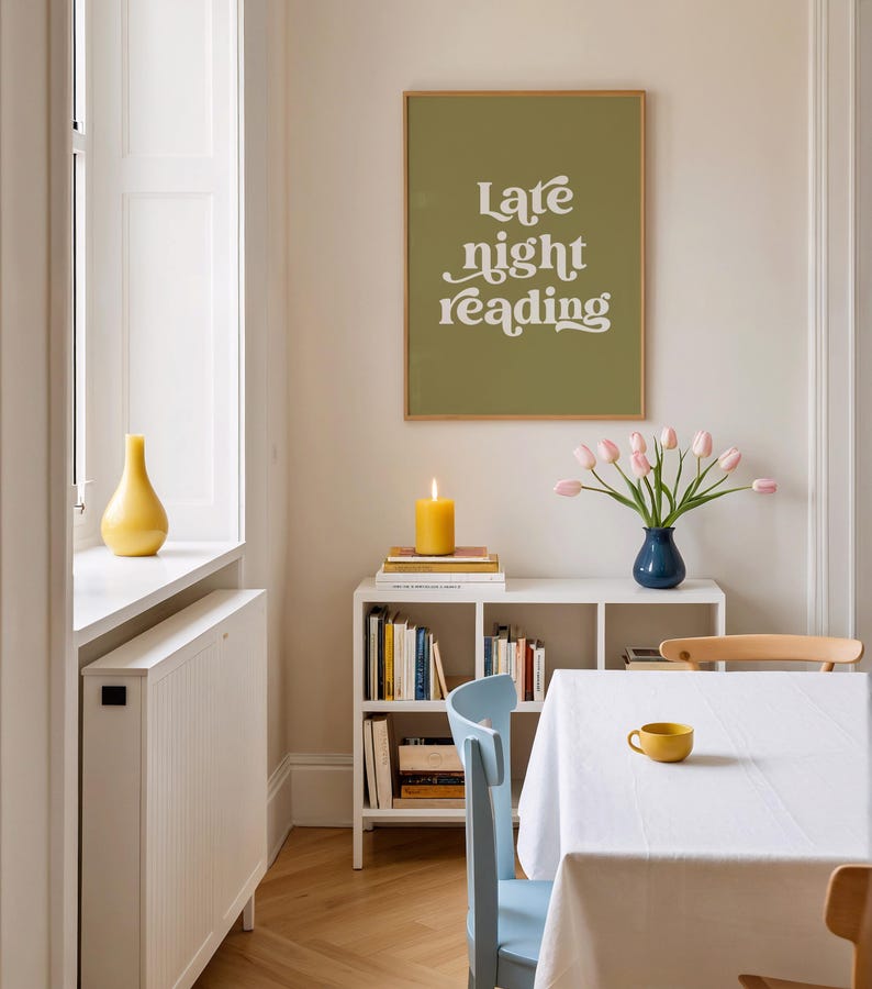 May include: A dining room scene featuring a framed print with the text "Late night reading" above a white shelf with books, a lit candle, and a blue vase of pink tulips. A yellow cup sits on a white tablecloth.
