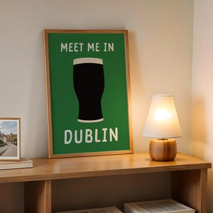 May include: A framed poster with the text "MEET ME IN DUBLIN" and a black pint glass silhouette on a green background. A small framed photo and a lamp with a white shade sit on a wooden shelf.