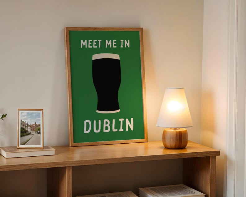 May include: A framed poster with the text "MEET ME IN DUBLIN" and a black pint glass silhouette on a green background. A small framed photo and a lamp with a white shade sit on a wooden shelf.
