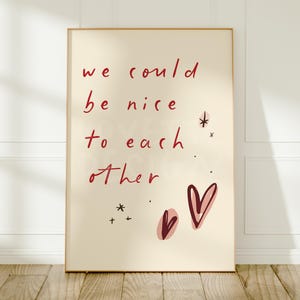 May include: A rectangular wall art print featuring a cream-coloured background with the red handwritten text "we could be nice to each other." The design includes two dark red heart illustrations and several small star-shaped accents scattered around.