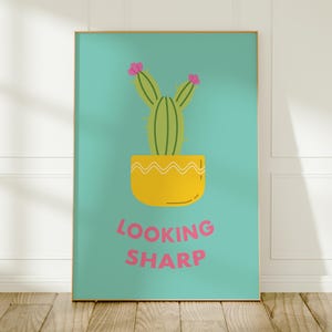 May include: A rectangular wall art print featuring a light teal background with a green cactus in a yellow pot. The cactus has two pink flowers on top. Below the pot, the text LOOKING SHARP is printed in pink capital letters in a bold font.