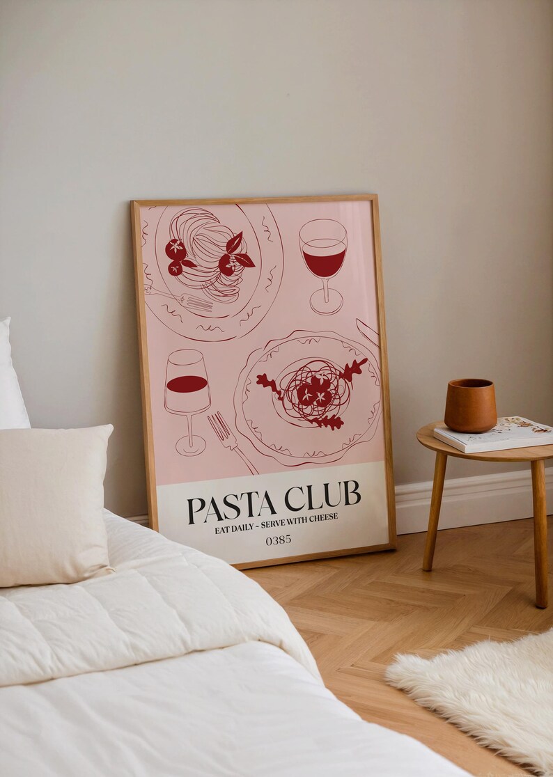 K&ouml;nnte beinhalten: Gerahmtes Poster mit rosa Hintergrund, das Strichzeichnungen von Pastagerichten und Weingl&auml;sern zeigt. Der Text "PASTA CLUB" steht unten, darunter "EAT DAILY - SERVE WITH CHEESE". Das Poster wird in einem Raum mit Bett und Beistelltisch angezeigt.