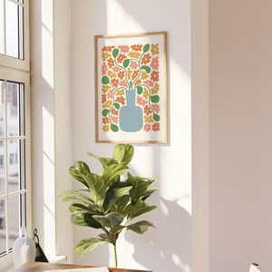 May include: A framed art print featuring a stylized floral arrangement. The artwork showcases a light blue vase filled with orange, yellow, and pink flowers with green stems and leaves. The print is set against a white background and framed in a light wood.