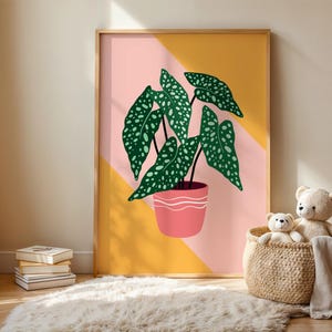 May include: A framed art print depicting a stylized illustration of a potted plant with large green leaves dotted with white spots. The plant is in a pink pot with a white wavy design, set against a pink and yellow geometric background.