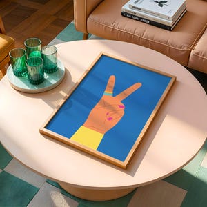 May include: A framed art print featuring a hand making the peace sign against a blue background. The hand is tan with a yellow sleeve, a blue ring, and pink nail polish. The artwork is displayed on a light pink round coffee table.