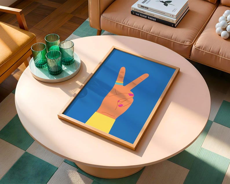 May include: A framed art print featuring a hand making the peace sign against a blue background. The hand is tan with a yellow sleeve, a blue ring, and pink nail polish. The artwork is displayed on a light pink round coffee table.