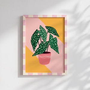 May include: A framed art print depicting a potted plant with large, green leaves dotted with white spots. The plant is in a pink pot, set against a pink and yellow geometric background. The frame has a pink and white striped pattern.