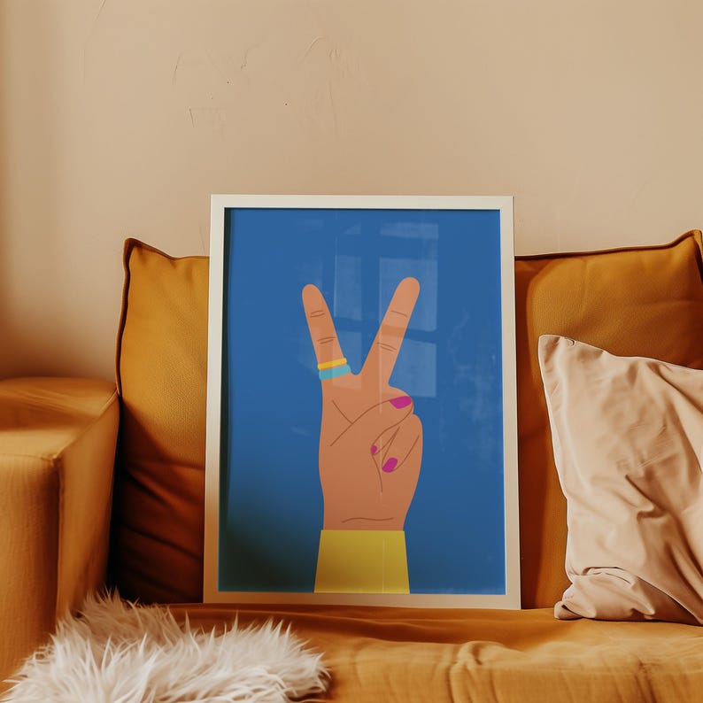 May include: A framed art print featuring a hand making the peace sign against a blue background. The hand has a gold and blue ring, pink fingernails, and a yellow sleeve. The artwork is displayed on a mustard-colored couch.