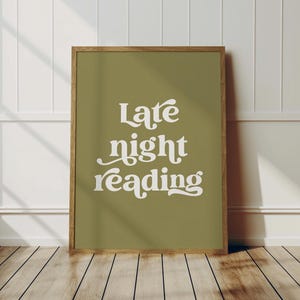 May include: A framed art print with the words "Late night reading" in white script on an olive green background. The wooden frame complements the color palette. The print is displayed against a white wall and wooden floor.