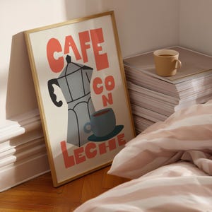 May include: A framed art print with a coffee theme. The print features a stylized illustration of a stovetop espresso maker, a coffee cup, and the words "CAFE CON LECHE" in coral-colored block letters. A beige mug sits on a stack of papers.