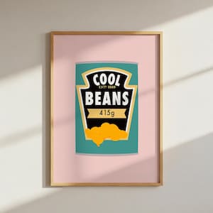 Puede incluir: Impresión enmarcada de una lata turquesa con la inscripción "COOL BEANS" en negro y dorado, y una ilustración de judías amarillas. La impresión está sobre un fondo rosa y enmarcada en un marco de madera marrón claro.
