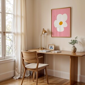 May include: A wooden desk with a matching chair, a gold desk lamp, and a framed print of a white flower with a yellow center on a pink background. A small vase with greenery and a framed photo are also on the desk.