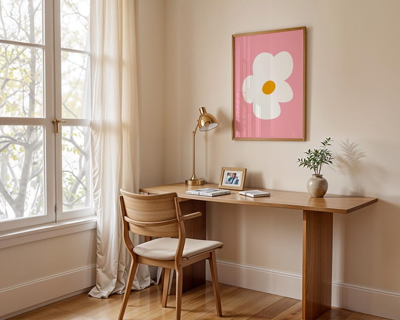 May include: A wooden desk with a matching chair, a gold desk lamp, and a framed print of a white flower with a yellow center on a pink background. A small vase with greenery and a framed photo are also on the desk.