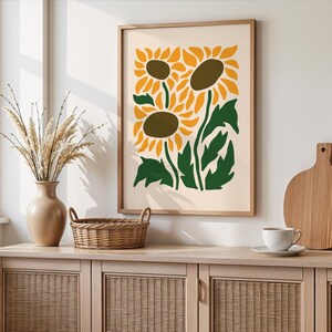 May include: A framed art print featuring a stylized illustration of sunflowers in yellow, green, and brown. The artwork is displayed on a light-colored wall above a wooden cabinet with a vase, basket, and other decorative items.