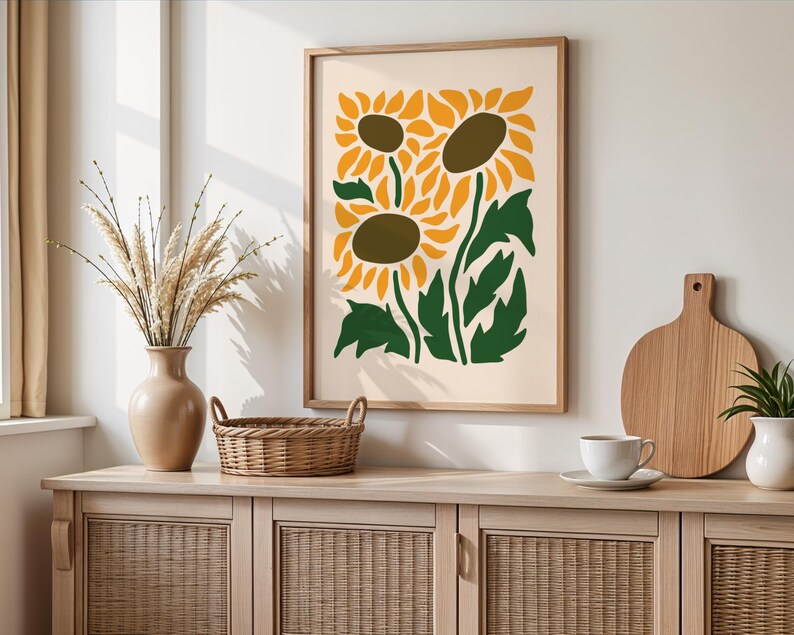 May include: A framed art print featuring a stylized illustration of sunflowers in yellow, green, and brown. The artwork is displayed on a light-colored wall above a wooden cabinet with a vase, basket, and other decorative items.