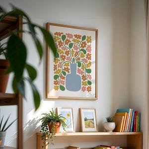 May include: A framed art print featuring a blue vase filled with colorful flowers in shades of pink, yellow, and orange. The artwork is displayed on a white wall, alongside other framed pictures and a wooden shelf with plants and books.