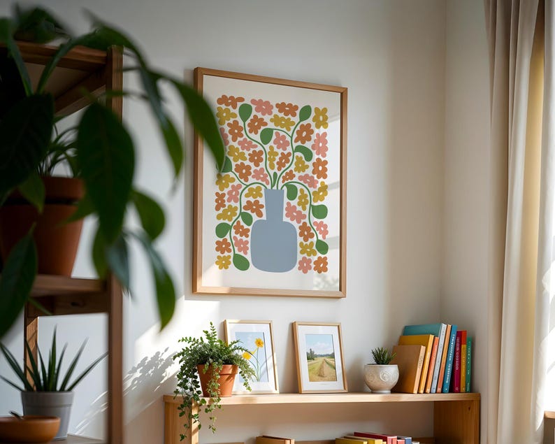 May include: A framed art print featuring a blue vase filled with colorful flowers in shades of pink, yellow, and orange. The artwork is displayed on a white wall, alongside other framed pictures and a wooden shelf with plants and books.