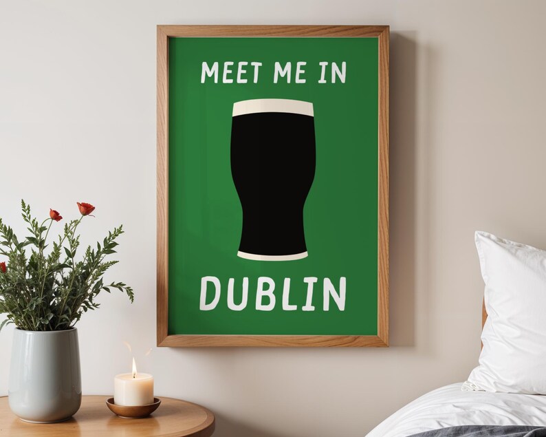 May include: A framed print with a green background and the words "MEET ME IN DUBLIN" in white. A black pint glass with a white rim is in the center. The frame is made of wood. A vase of flowers and a lit candle are in the foreground.