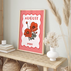 May include: A framed art print featuring a floral illustration of poppies. The print has a red border with the word "AUGUST" at the top and "poppy" at the bottom. The artwork is displayed on a light-colored wooden bench.