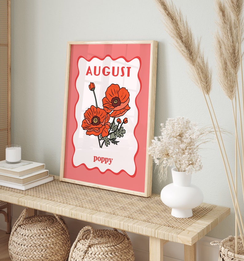 May include: A framed art print featuring a floral illustration of poppies. The print has a red border with the word "AUGUST" at the top and "poppy" at the bottom. The artwork is displayed on a light-colored wooden bench.