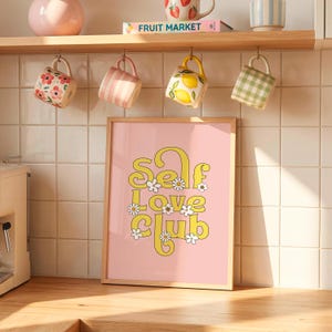 May include: A framed vertical wall art print featuring a light pink background with the yellow text "Self Love Club" in a retro font, decorated with small white daisy flowers. The print is displayed on a kitchen counter against a tiled wall.