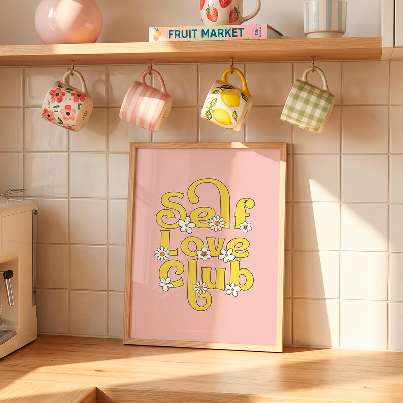 May include: A framed vertical wall art print featuring a light pink background with the yellow text "Self Love Club" in a retro font, decorated with small white daisy flowers. The print is displayed on a kitchen counter against a tiled wall.