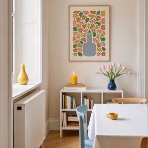 May include: A dining room scene with a framed floral print, a white bookshelf, and a table set for a meal. The print features a vase with colorful flowers. A yellow candle and a vase of pink tulips add color to the room. A yellow cup sits on the table.