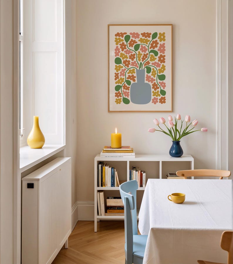 May include: A dining room scene with a framed floral print, a white bookshelf, and a table set for a meal. The print features a vase with colorful flowers. A yellow candle and a vase of pink tulips add color to the room. A yellow cup sits on the table.