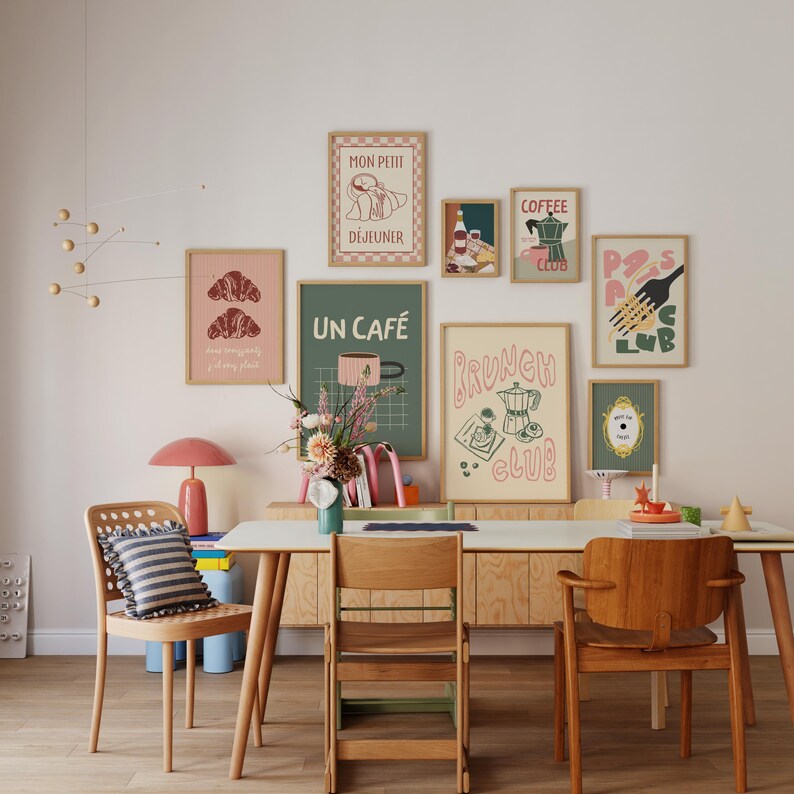 May include: A collection of framed art prints with a cafe theme, including images of croissants, coffee, and brunch. The prints are in various sizes and colors, with a mix of text and illustrations. The room also features wooden chairs, a table, and a pink lamp.