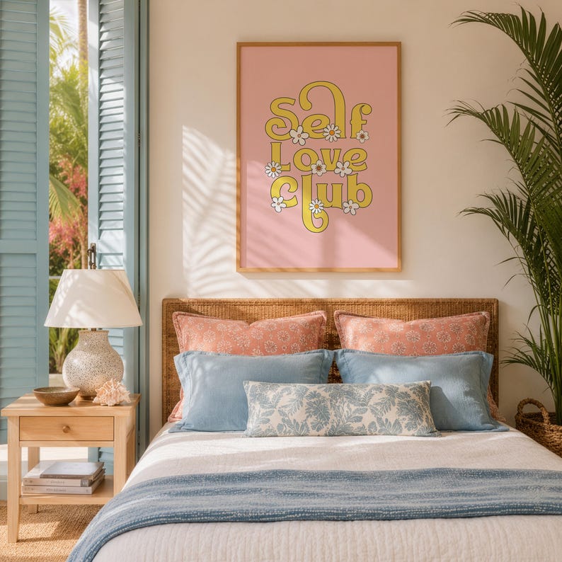 May include: A rectangular wall art print in a light wood frame featuring the yellow text Self Love Club with white daisy accents on a solid pink background, displayed above a bed with coral and blue pillows and a white textured bedspread.
