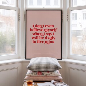 K&ouml;nnte beinhalten: Ein gerahmter Kunstdruck mit hellrosa Hintergrund und rotem Text, der lautet: I don't even believe myself when I say I will be ready in five mins. Der Druck befindet sich in einem d&uuml;nnen schwarzen Rahmen &uuml;ber einem Stapel von drei Kissen.