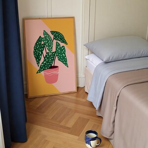 May include: A framed botanical print of a green plant with white spots in a pink pot, set against a yellow and pink geometric background. A bed with grey and beige bedding and a blue and white patterned mug are also visible.