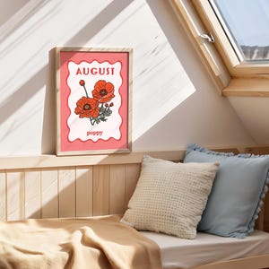 May include: A framed art print featuring a poppy flower illustration. The artwork has a pink background with the word "August" at the top and "poppy" at the bottom. The frame is made of light-colored wood.