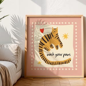 K&ouml;nnte beinhalten: Ein quadratischer Wandkunstdruck in einem Rahmen, der einen stilisierten orangefarbenen Tiger mit schwarzen Streifen auf cremefarbenem Hintergrund zeigt. Das Bild enth&auml;lt ein kleines rotes Herz, zwei gelbe Sterne und den schwarzen Text "wash your paws".