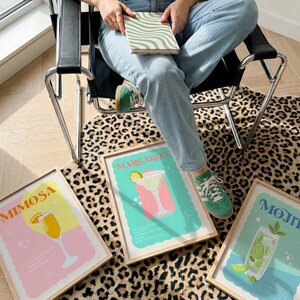 May include: Three framed cocktail art prints: "Mimosa," "Margarita," and "Mojito." Each print features a colorful illustration of the drink with its name in a retro font. The prints are displayed on a leopard print rug.
