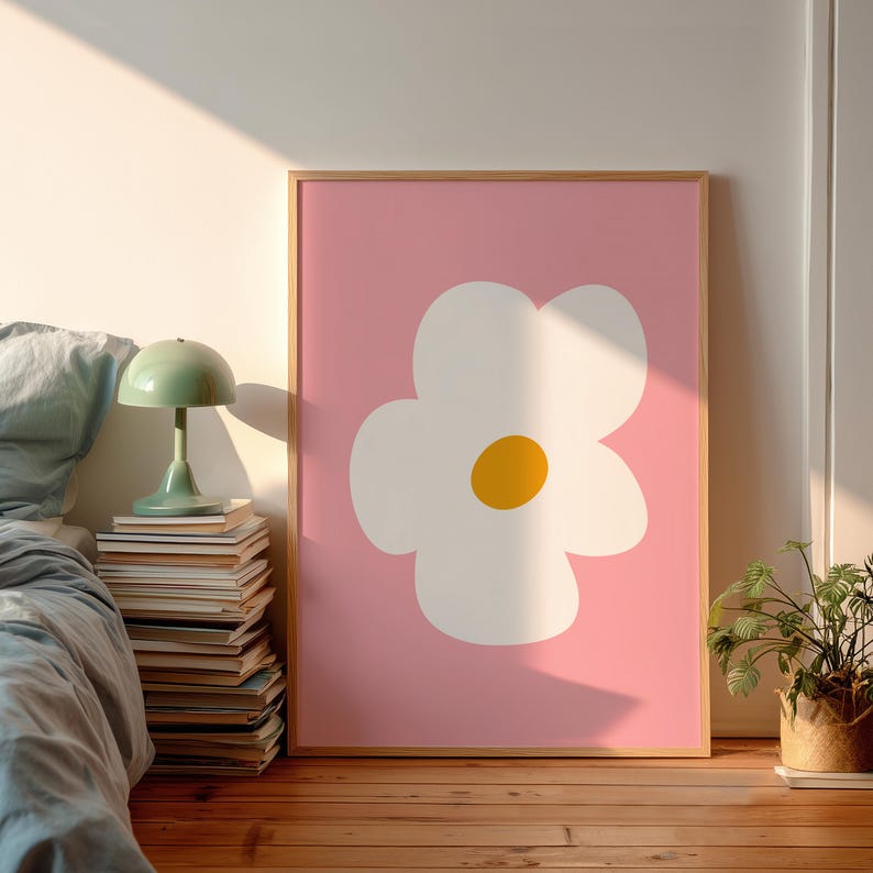 May include: A framed art print featuring a large white flower with a yellow center against a pink background. The artwork is in a wooden frame and is displayed in a room with a bed, a lamp, and a small potted plant.