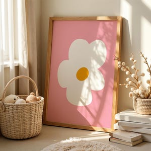 May include: A framed art print featuring a large white flower with a yellow center on a pink background. The wooden frame leans against a wall. A woven basket with stuffed toys sits on the floor, near a rug and a window.