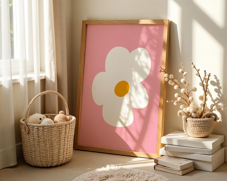 May include: A framed art print featuring a large white flower with a yellow center on a pink background. The wooden frame leans against a wall. A woven basket with stuffed toys sits on the floor, near a rug and a window.