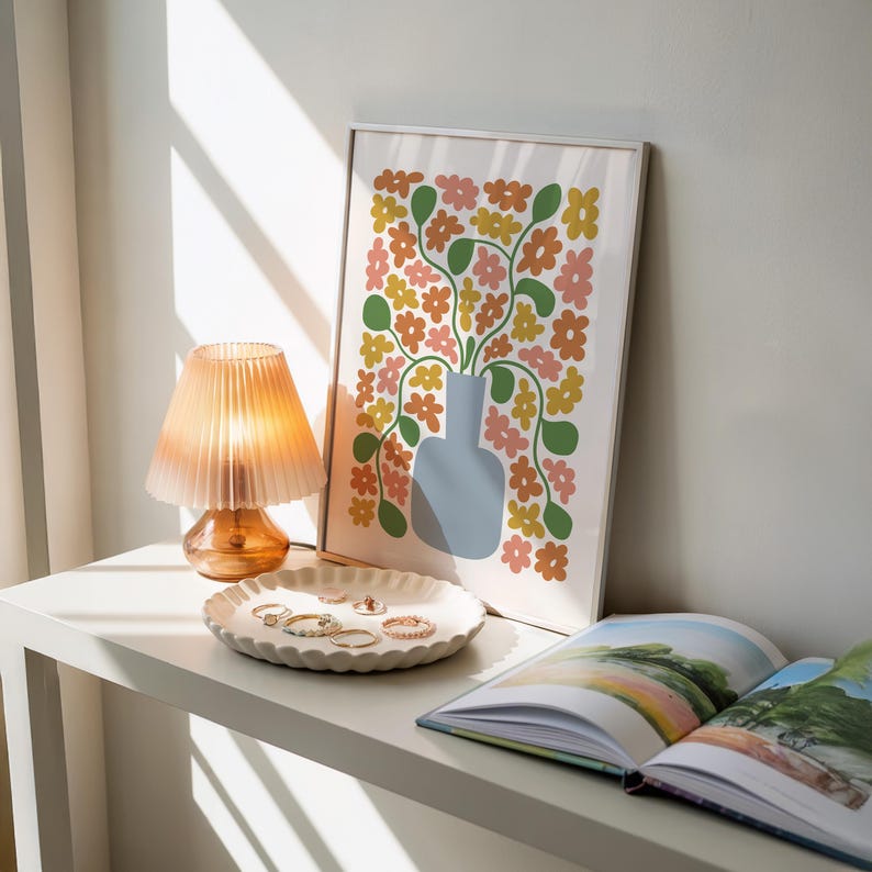 May include: A decorative arrangement on a white table. A small, orange glass lamp with a pleated shade sits next to a white, scalloped dish holding rings. A framed floral print and an open book complete the scene. The print features a blue vase with colorful flowers.