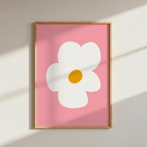 May include: A framed art print featuring a large, stylized white flower with a yellow center against a pink background. The artwork is in a wooden frame and is displayed on a white wall. The design is simple and modern.