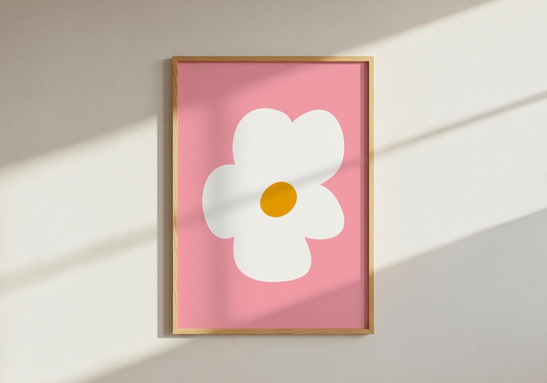 May include: A framed art print featuring a large, stylized white flower with a yellow center against a pink background. The artwork is in a wooden frame and is displayed on a white wall. The design is simple and modern.