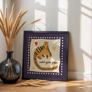 K&ouml;nnte beinhalten: Ein quadratischer Kunstdruck in einem Rahmen zeigt eine stilisierte orangefarbene Katze mit dunklen Streifen, ein kleines rotes Herz und gelbe Sterne. Der Text 'wash your paws' ist in Schwarz gedruckt. Der Rand ist dunkelviolett mit wei&szlig;en Punkten.