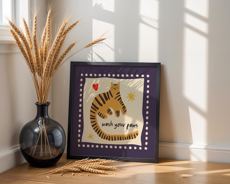 K&ouml;nnte beinhalten: Ein quadratischer Kunstdruck in einem Rahmen zeigt eine stilisierte orangefarbene Katze mit dunklen Streifen, ein kleines rotes Herz und gelbe Sterne. Der Text 'wash your paws' ist in Schwarz gedruckt. Der Rand ist dunkelviolett mit wei&szlig;en Punkten.