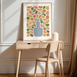 May include: A framed floral art print featuring a blue vase filled with colorful flowers in shades of orange, pink, and yellow. The artwork is displayed on a light wood desk with a matching chair, creating a modern and stylish home decor scene.